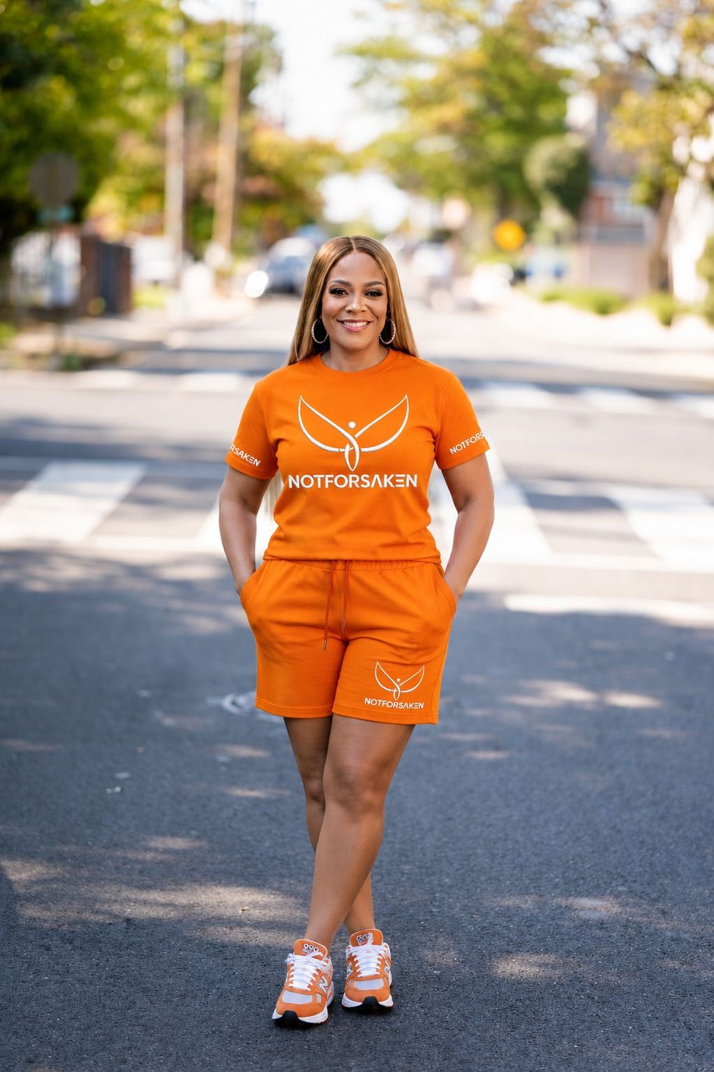 Smiling woman in an orange Notforsaken outfit stands on a city street with hands in pockets and crosswalk behind her.